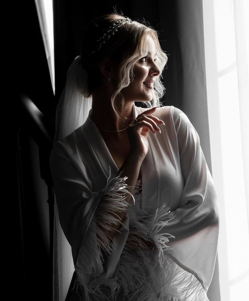 May include: A bride in a white satin robe with feather trim, standing by a window. She is wearing a veil and a tiara.