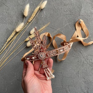 May include: A rose gold headband with a delicate floral design and pearl accents. The headband is adorned with sparkling rhinestones and features a dangling pearl drop.
