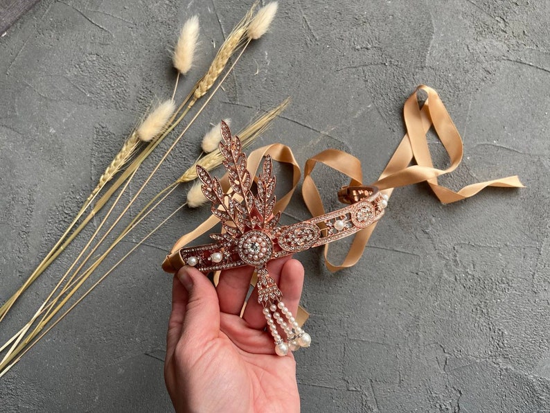 May include: A rose gold headband with a delicate floral design and pearl accents. The headband is adorned with sparkling rhinestones and features a dangling pearl drop.