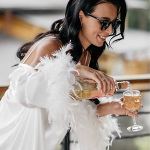 May include: A woman in a white satin robe with feather trim pours champagne into a glass. She is wearing sunglasses and smiling.