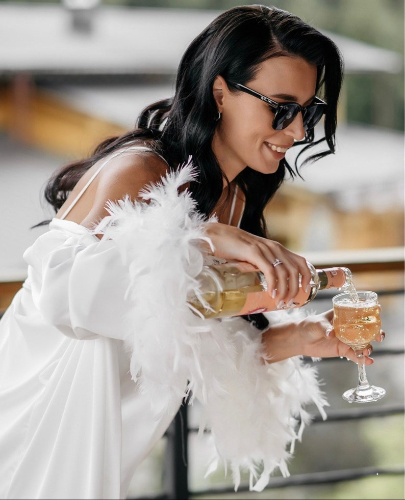 May include: A woman in a white satin robe with feather trim pours champagne into a glass. She is wearing sunglasses and smiling.