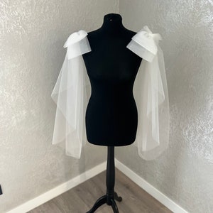 May include: White tulle bridal bolero with large bows on the shoulders. The bolero is displayed on a black dress form.