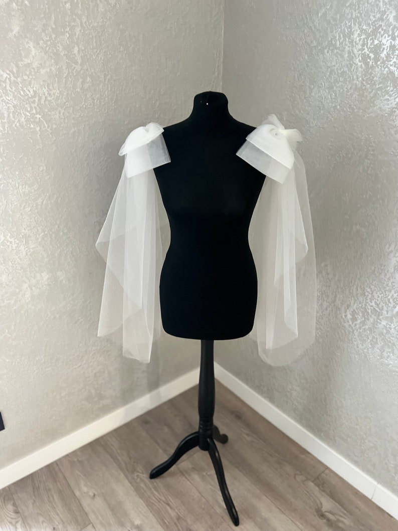 May include: White tulle bridal bolero with large bows on the shoulders. The bolero is displayed on a black dress form.
