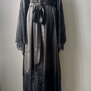 K&ouml;nnte beinhalten: Eine schwarze Satin-Robe mit langen &Auml;rmeln und einem Spitzenbesatz. Die Robe hat einen Bindeg&uuml;rtel und einen V-Ausschnitt.