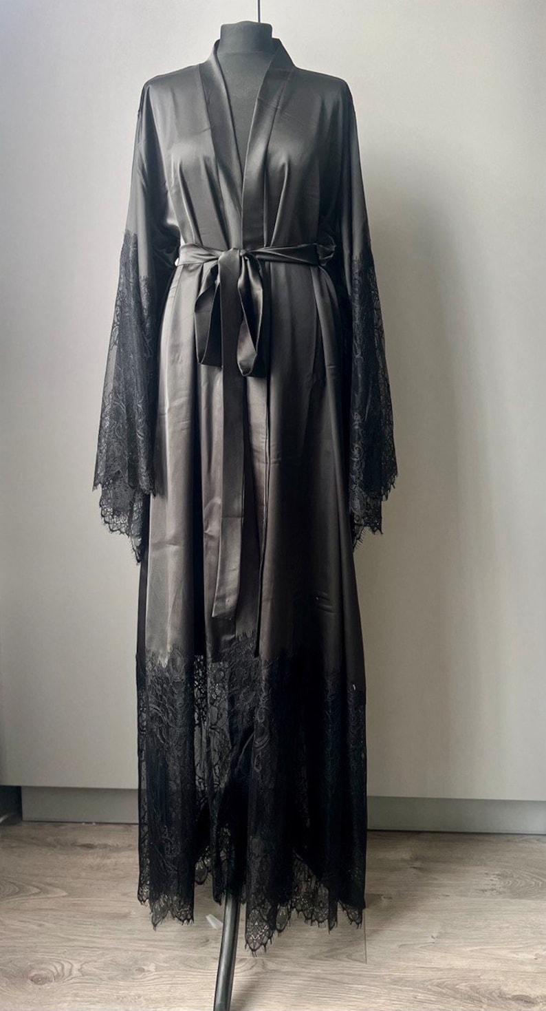 K&ouml;nnte beinhalten: Eine schwarze Satin-Robe mit langen &Auml;rmeln und einem Spitzenbesatz. Die Robe hat einen Bindeg&uuml;rtel und einen V-Ausschnitt.