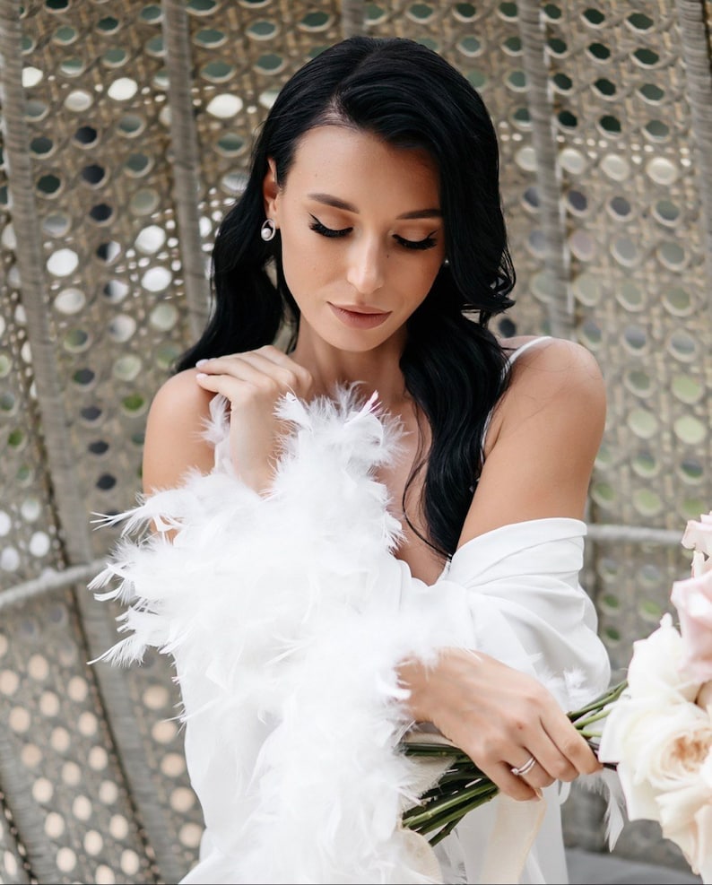 May include: A woman in a white robe with a feather trim holds a bouquet of white and pink roses. She is wearing a pearl earring and a ring on her finger.