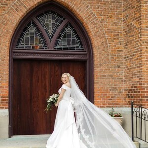 May include: A bride in a white off-the-shoulder wedding dress with a long veil, holding a bouquet of white flowers, stands in front of a brick building with a wooden door.