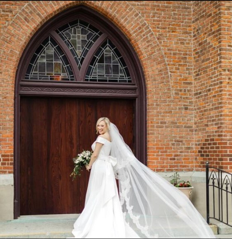 May include: A bride in a white off-the-shoulder wedding dress with a long veil, holding a bouquet of white flowers, stands in front of a brick building with a wooden door.