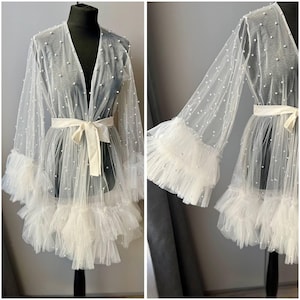 May include: A white sheer robe with pearl embellishments and a ruffled trim. The robe has a tie at the waist and long sleeves with ruffled cuffs.