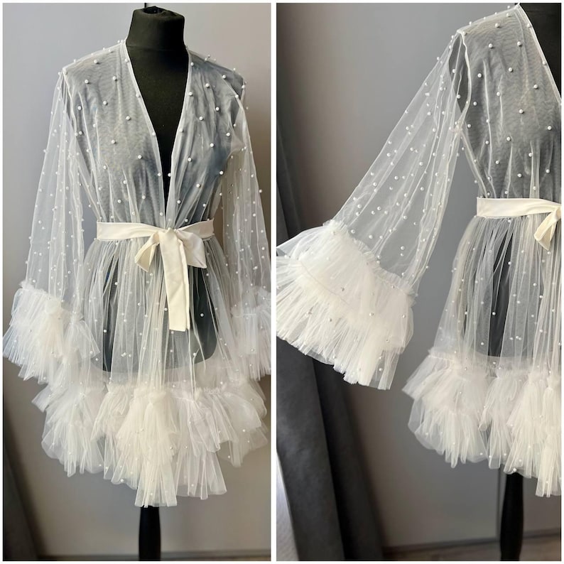May include: A white sheer robe with pearl embellishments and a ruffled trim. The robe has a tie at the waist and long sleeves with ruffled cuffs.