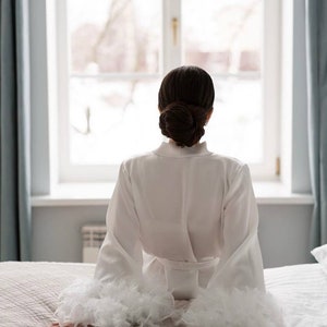 May include: A white satin robe with feather trim on the sleeves. The robe is tied at the waist and the person wearing it is sitting on a bed looking out a window.