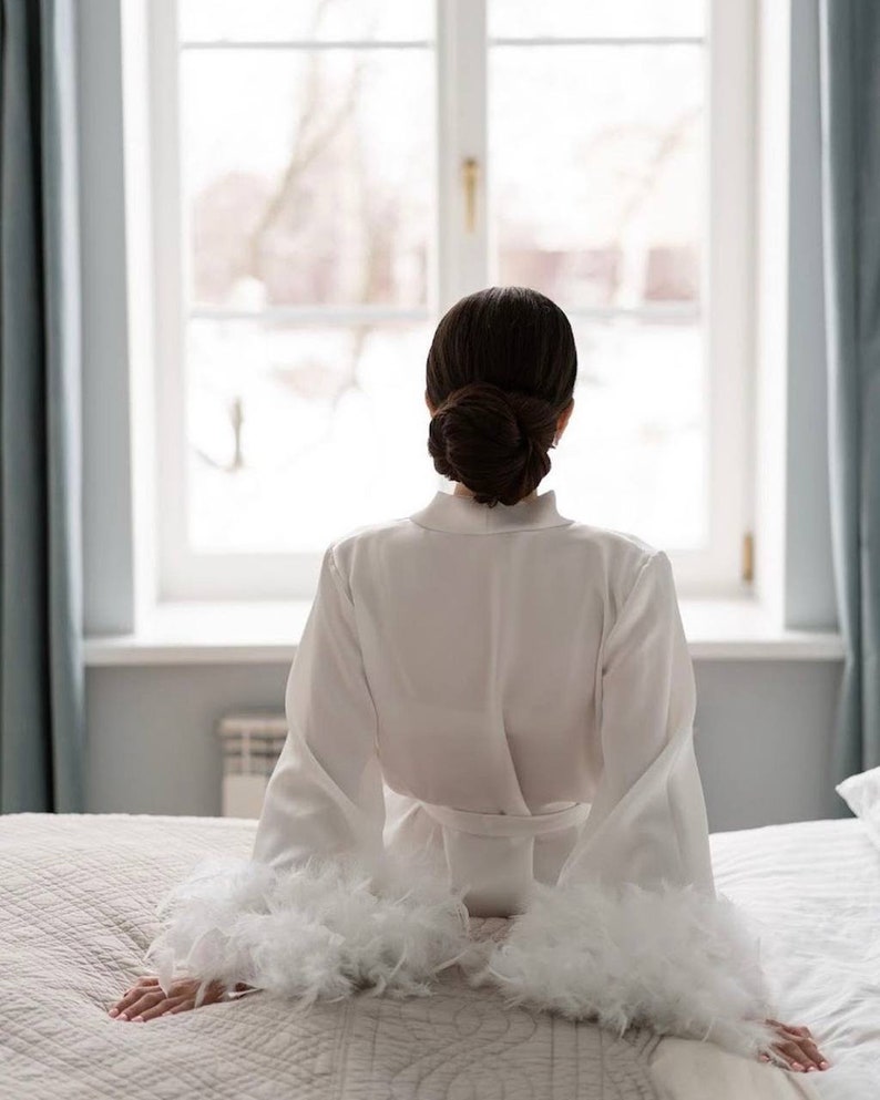 May include: A white satin robe with feather trim on the sleeves. The robe is tied at the waist and the person wearing it is sitting on a bed looking out a window.