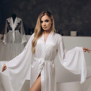 May include: A white satin robe with long sleeves and a tie belt. The robe has a wide, flowing silhouette and a delicate lace trim at the cuffs and hem.