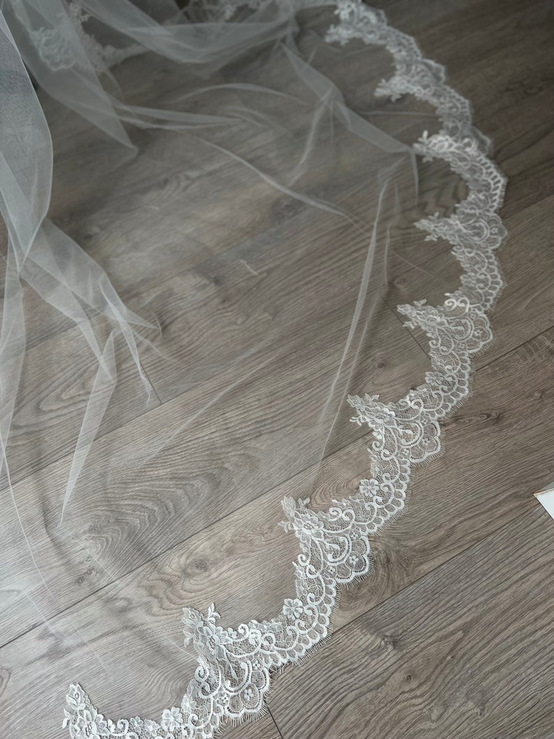 May include: A white tulle wedding veil with a delicate lace trim. The lace trim features a floral design with intricate details.