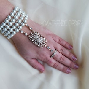 May include: A gold hand chain with a large, oval-shaped pendant adorned with rhinestones. The chain is attached to a bracelet made of multiple strands of white pearls. The bracelet and chain are worn on a hand with a ring on the middle finger.