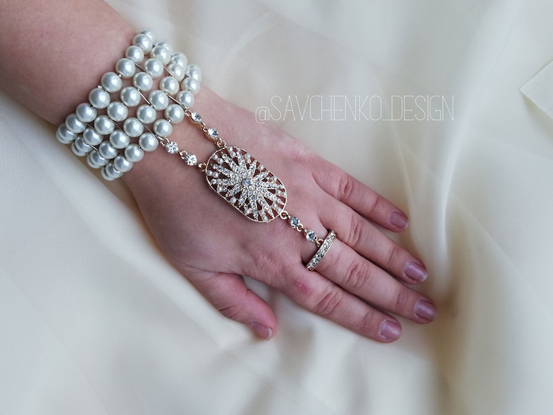 May include: A gold hand chain with a large, oval-shaped pendant adorned with rhinestones. The chain is attached to a bracelet made of multiple strands of white pearls. The bracelet and chain are worn on a hand with a ring on the middle finger.
