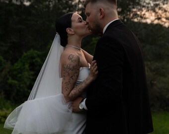 May include: A bride and groom kiss in a forest setting. The bride is wearing a white wedding dress with a veil and a floral tattoo on her arm. The groom is wearing a black suit.