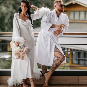 May include: A couple wearing white robes, the woman holding a bouquet of pink flowers. The woman's robe has feather trim. The man is wearing sunglasses.
