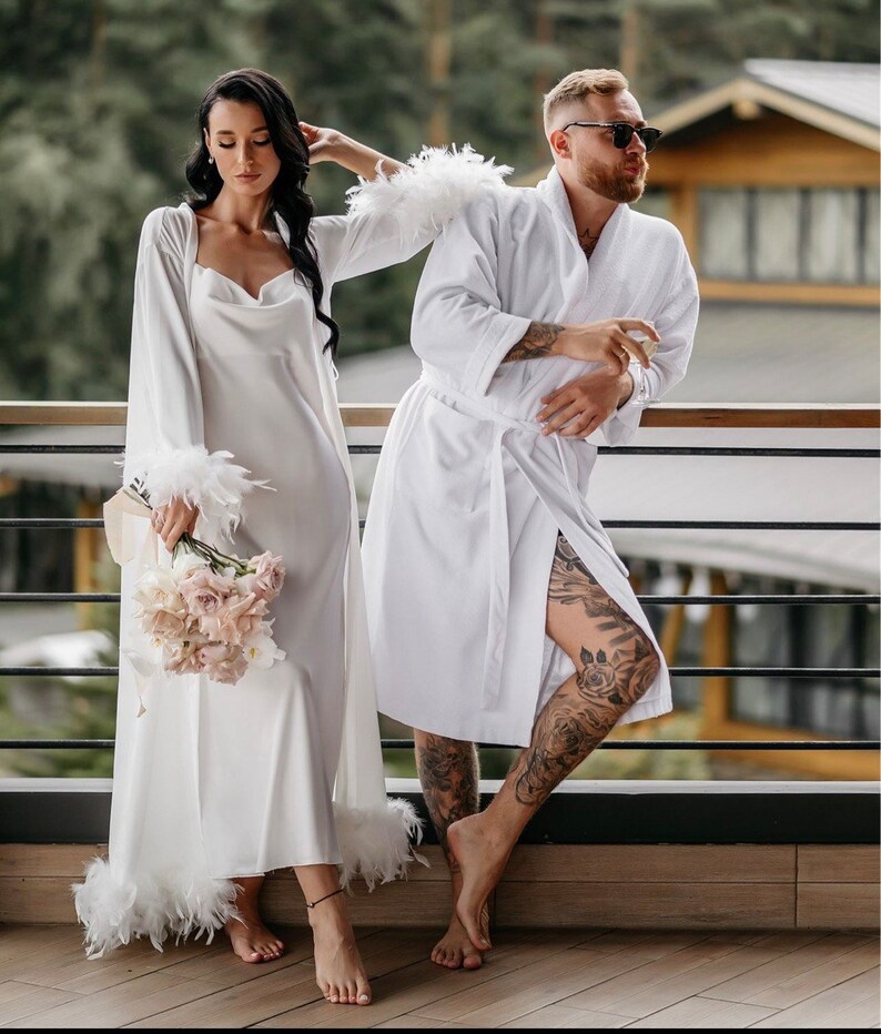 May include: A couple wearing white robes, the woman holding a bouquet of pink flowers. The woman's robe has feather trim. The man is wearing sunglasses.