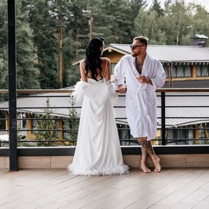 May include: A couple in white robes stand on a balcony overlooking a forest. The woman is wearing a white robe with feather trim and the man is wearing a white robe with a black belt. They are both looking at each other.