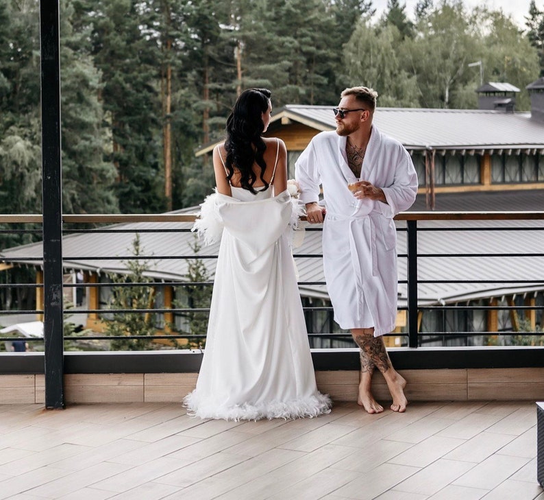 May include: A couple in white robes stand on a balcony overlooking a forest. The woman is wearing a white robe with feather trim and the man is wearing a white robe with a black belt. They are both looking at each other.
