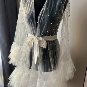 May include: A white sheer robe with pearl embellishments and a satin sash. The robe has a ruffled hem and long sleeves.