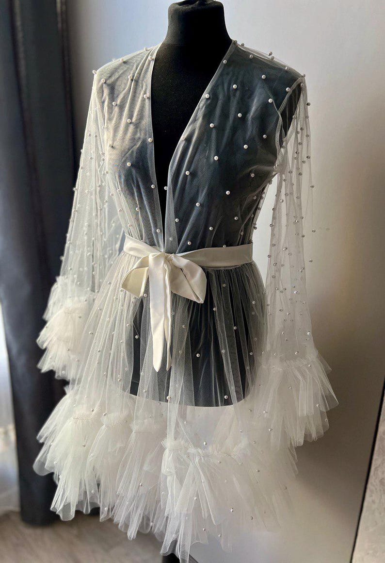 May include: A white sheer robe with pearl embellishments and a satin sash. The robe has a ruffled hem and long sleeves.