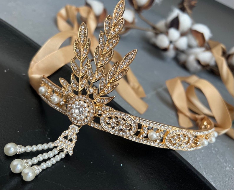 May include: A gold headband with a delicate design of leaves and rhinestones. The headband is adorned with small white pearls and has a dangling pearl tassel.