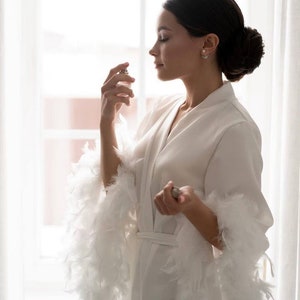 May include: A woman in a white robe with feather trim sprays perfume. The robe has a sash and long sleeves. The woman is standing in front of a window.