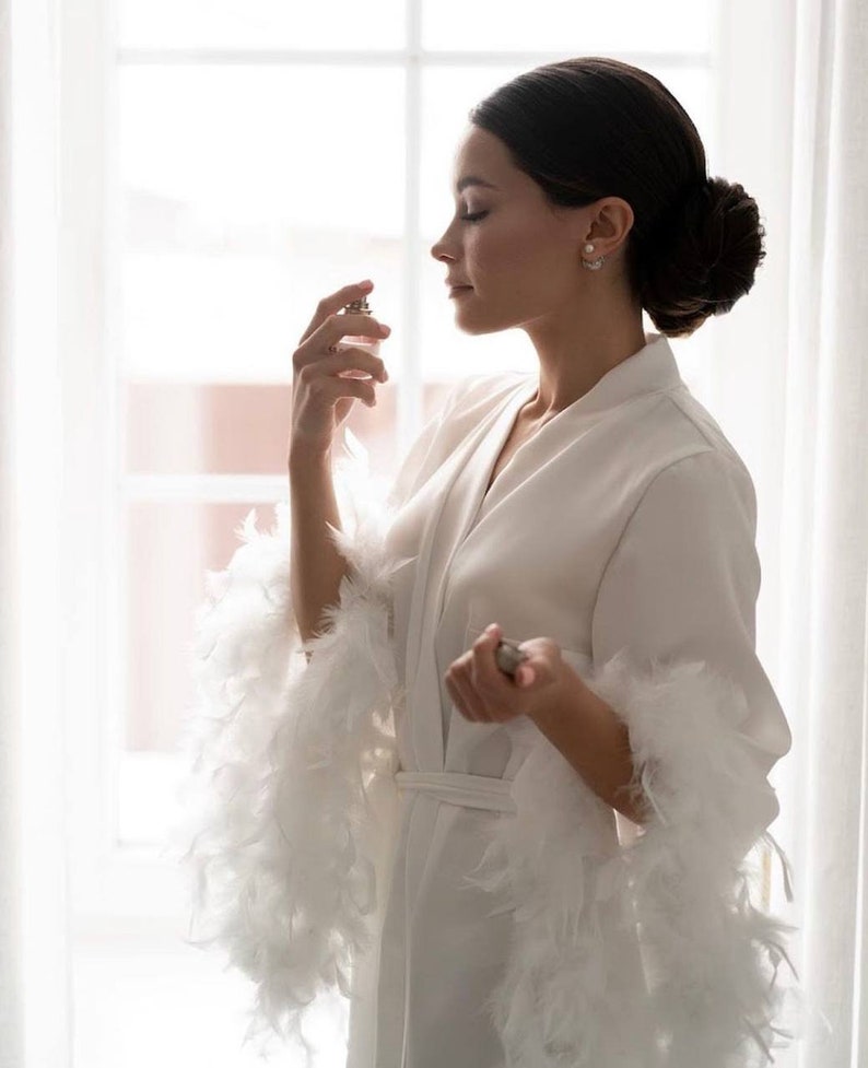 May include: A woman in a white robe with feather trim sprays perfume. The robe has a sash and long sleeves. The woman is standing in front of a window.