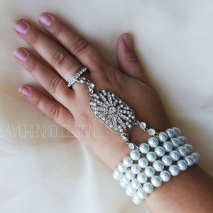May include: A silver ring with a crystal design and a pearl bracelet with a silver charm. The bracelet is worn on a hand.