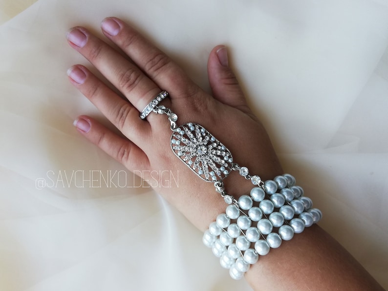 May include: A silver ring with a crystal design and a pearl bracelet with a silver charm. The bracelet is worn on a hand.