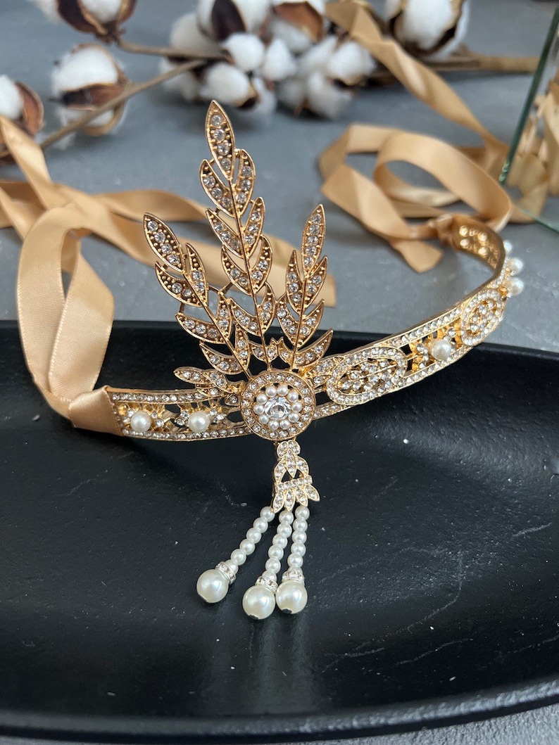 May include: A gold headband with a pearl and rhinestone design. The headband features a leaf motif and a dangling pearl tassel.