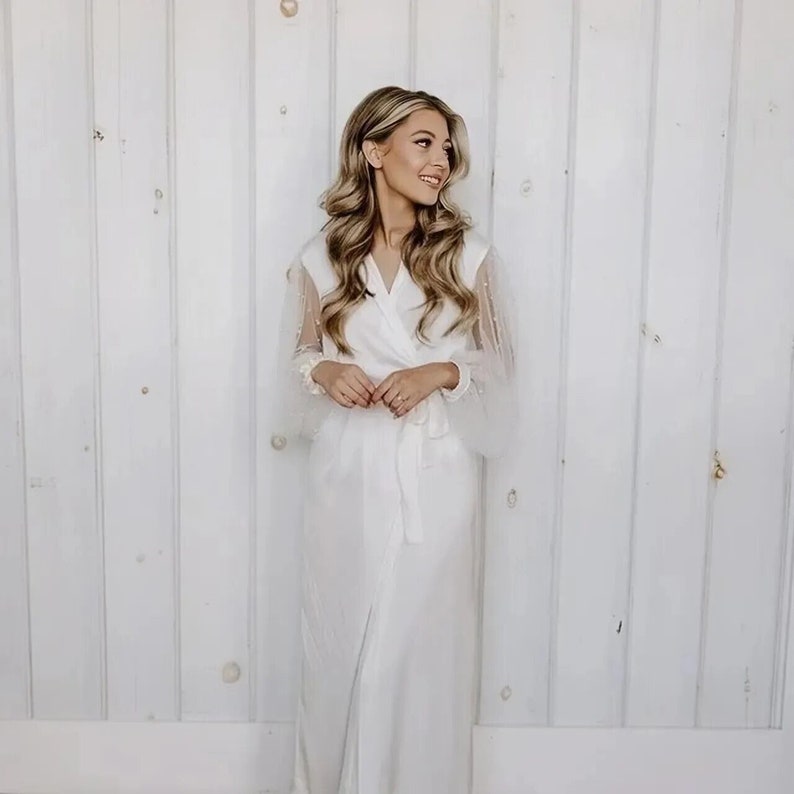 May include: A white satin robe with long sleeves and a sheer, beaded overlay. The robe has a wrap closure and a sash tie at the waist.