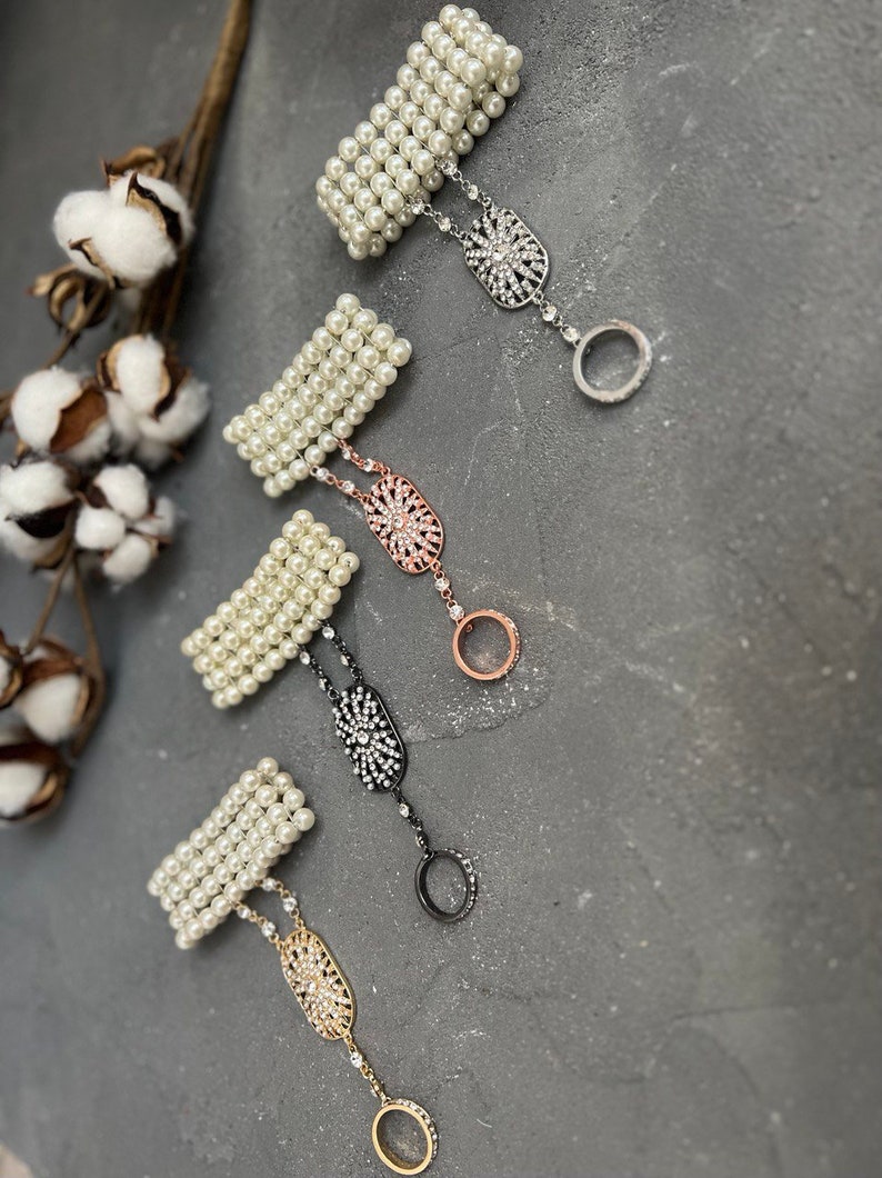 May include: Four pearl bracelets with a rhinestone ring attached to each. The bracelets are white and the rings are silver, rose gold, black, and gold.