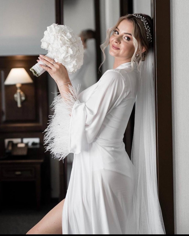 May include: A bride in a white satin robe with feather trim and a white veil, holding a white hydrangea bouquet. She is wearing a silver tiara.