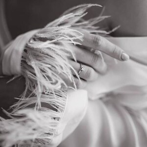 May include: A close-up of a hand wearing a ring with a small stone, the hand is wearing a white garment with a feather trim.