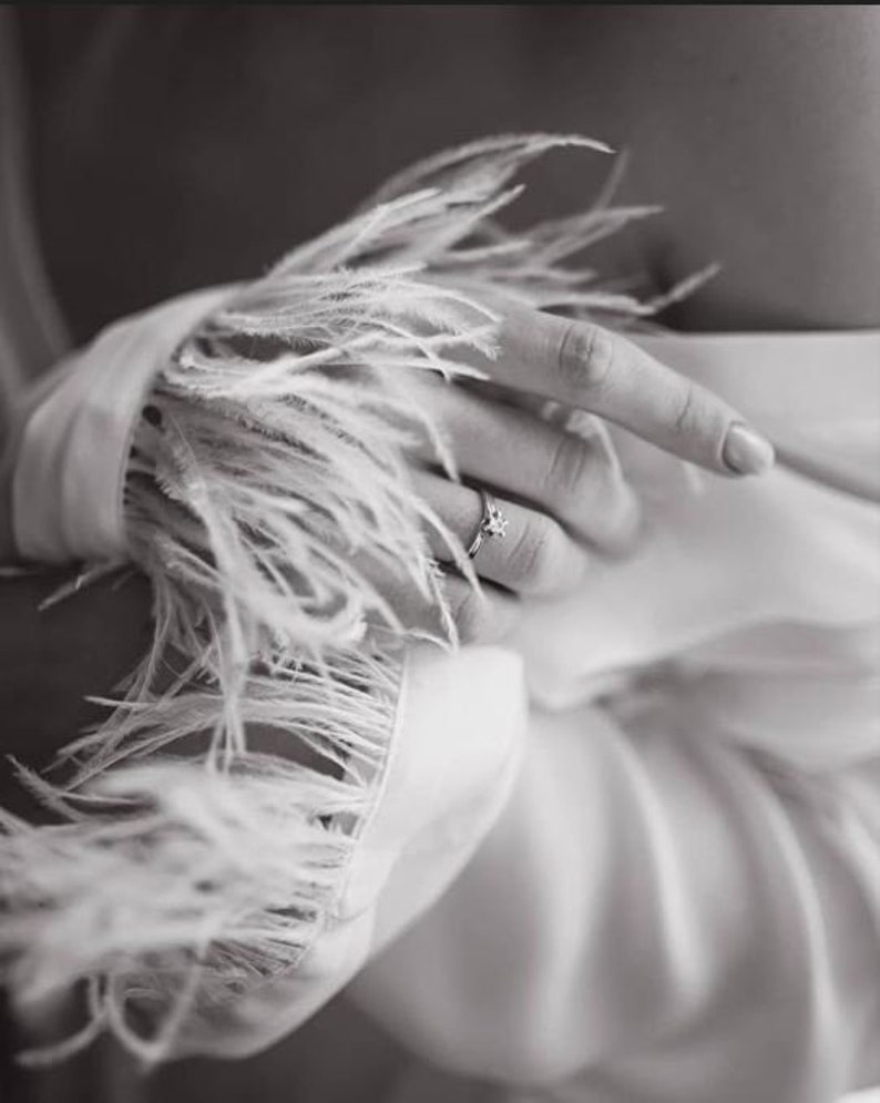 May include: A close-up of a hand wearing a ring with a small stone, the hand is wearing a white garment with a feather trim.