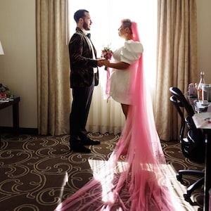 Könnte beinhalten: Ein Paar steht in einem Hotelzimmer, die Frau trägt ein weißes Kleid und einen langen rosa Schleier, der Mann trägt eine braune gemusterte Sakkojacke. Die Frau hält einen Blumenstrauß.