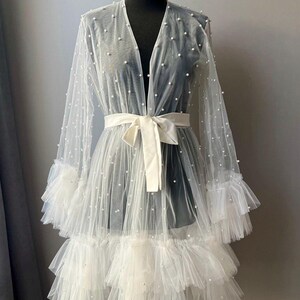 May include: A white sheer robe with a ruffled hem and pearl embellishments. The robe has a V-neckline and a sash tie at the waist.