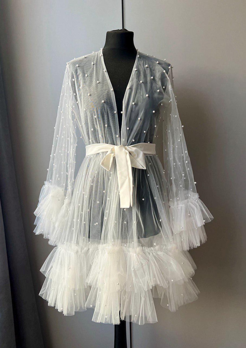 May include: A white sheer robe with a ruffled hem and pearl embellishments. The robe has a V-neckline and a sash tie at the waist.