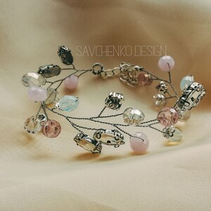May include: A delicate silver wire bracelet adorned with pastel pink, blue, and clear glass beads. The bracelet features a unique design with branches and leaves, creating a whimsical and elegant look. The text "SAVCHENKO DESIGN" is visible on the bracelet.