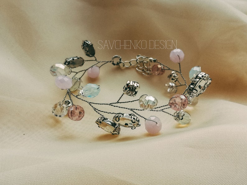 May include: A delicate silver wire bracelet adorned with pastel pink, blue, and clear glass beads. The bracelet features a unique design with branches and leaves, creating a whimsical and elegant look. The text "SAVCHENKO DESIGN" is visible on the bracelet.