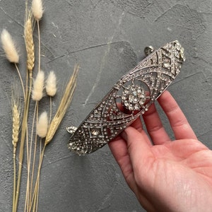 May include: A silver tiara with a geometric design and clear rhinestones. The tiara is on a gray background.