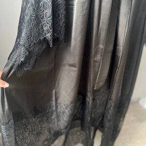 K&ouml;nnte beinhalten: Schwarze Satinrobe mit Spitzenbesatz. Die Robe hat eine lange, flie&szlig;ende Silhouette und einen Bindeg&uuml;rtel.