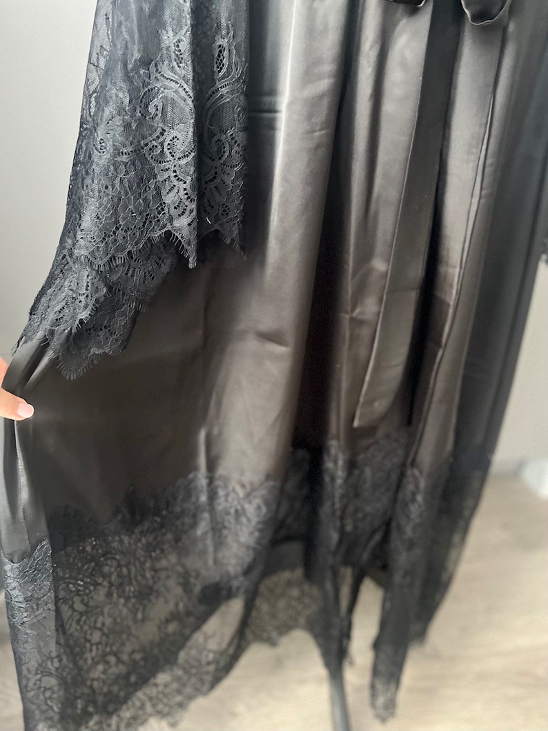 K&ouml;nnte beinhalten: Schwarze Satinrobe mit Spitzenbesatz. Die Robe hat eine lange, flie&szlig;ende Silhouette und einen Bindeg&uuml;rtel.