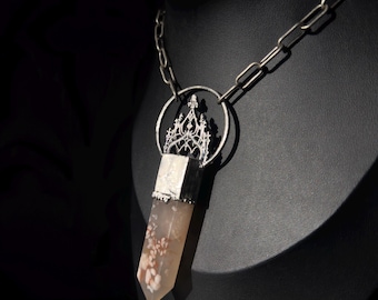 May include: A silver-toned necklace with a pendant featuring a crystal point. The crystal is a light peach color with white floral inclusions. The pendant is attached to a silver chain with a decorative circular element.