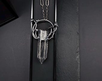 May include: A silver necklace with a crystal pendant. The pendant is shaped like a long, pointed crystal and is surrounded by a silver frame. The necklace is on a black background.