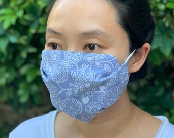 Puede incluir: Mascarilla facial gris azulada con un estampado floral blanco. La mascarilla es de tela y tiene gomas elásticas blancas para las orejas. El texto "Gray Blue" está en la parte inferior izquierda.