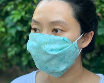 Puede incluir: Una persona lleva una mascarilla facial azul turquesa con un estampado floral blanco. La mascarilla está hecha de tela y tiene una banda elástica blanca. El texto "Vine Teal" y "DIAMOND CITY CO." es visible en la imagen.